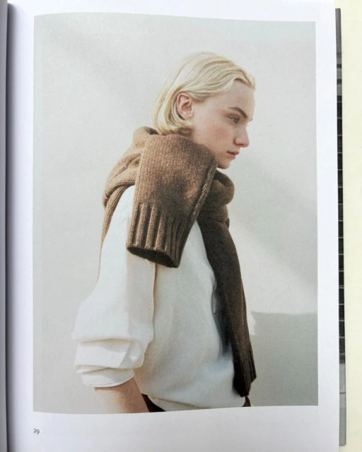 Blonde adult poses in a soft light wearing a white long sleeve shirt and a brown knit scarf draped over the shoulders and chest.
