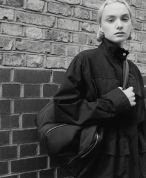 Model Mollie Cotter in a dark high-collar jacket and shoulder bag posing before a brick wall in black and white.