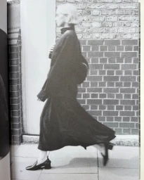 A woman in a long black dress and low-heeled black shoes walks briskly in profile past a brick wall on an outdoor pavement.