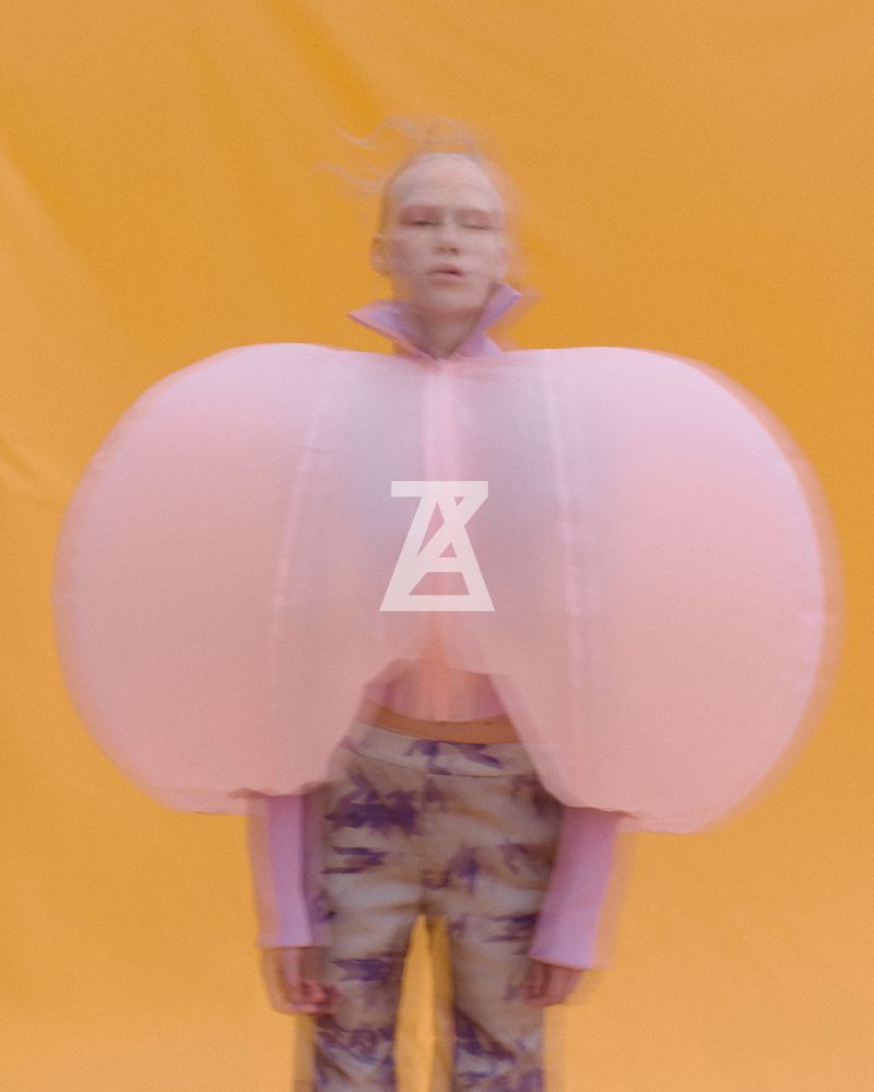 A person with light skin and hair poses in a unique fashion outfit with large pink arm pieces against a bright yellow background.