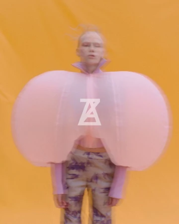 A person with light skin and hair poses in a unique fashion outfit with large pink arm pieces against a bright yellow background.