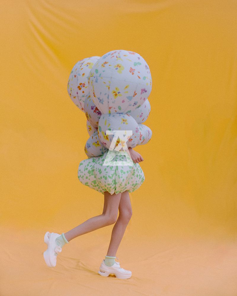 A person walks in a floral print dress and sneakers against a yellow background, captured in a fashion-forward studio shot.