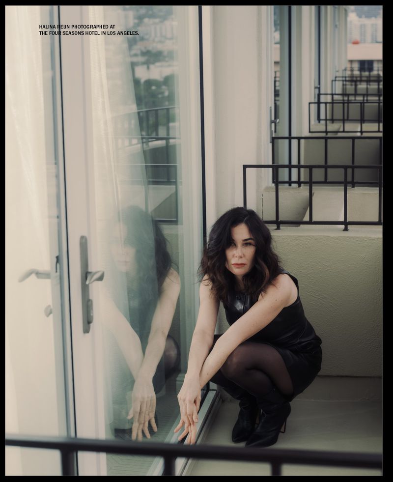 A dark-haired woman in a black dress poses crouching on a balcony with a glass door and ornate railing, outside at the Four Seasons.