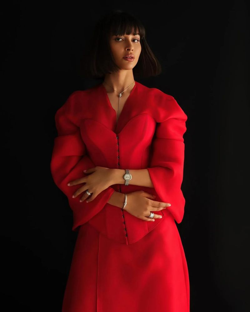 Portrait of a woman with dark bob hairstyle in a striking red two-piece outfit against a stark black backdrop, jewelry on her hands and neck.