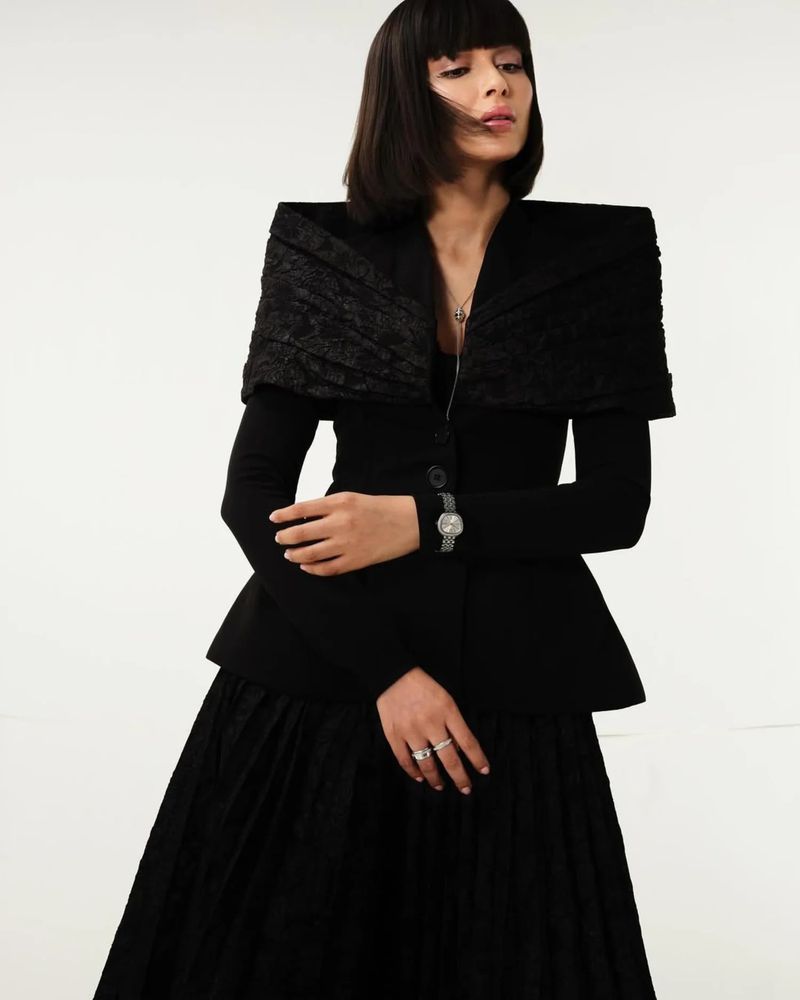 Elegant adult female in a black textured dress and jacket in a studio setting, posing gracefully with a serious facial expression.