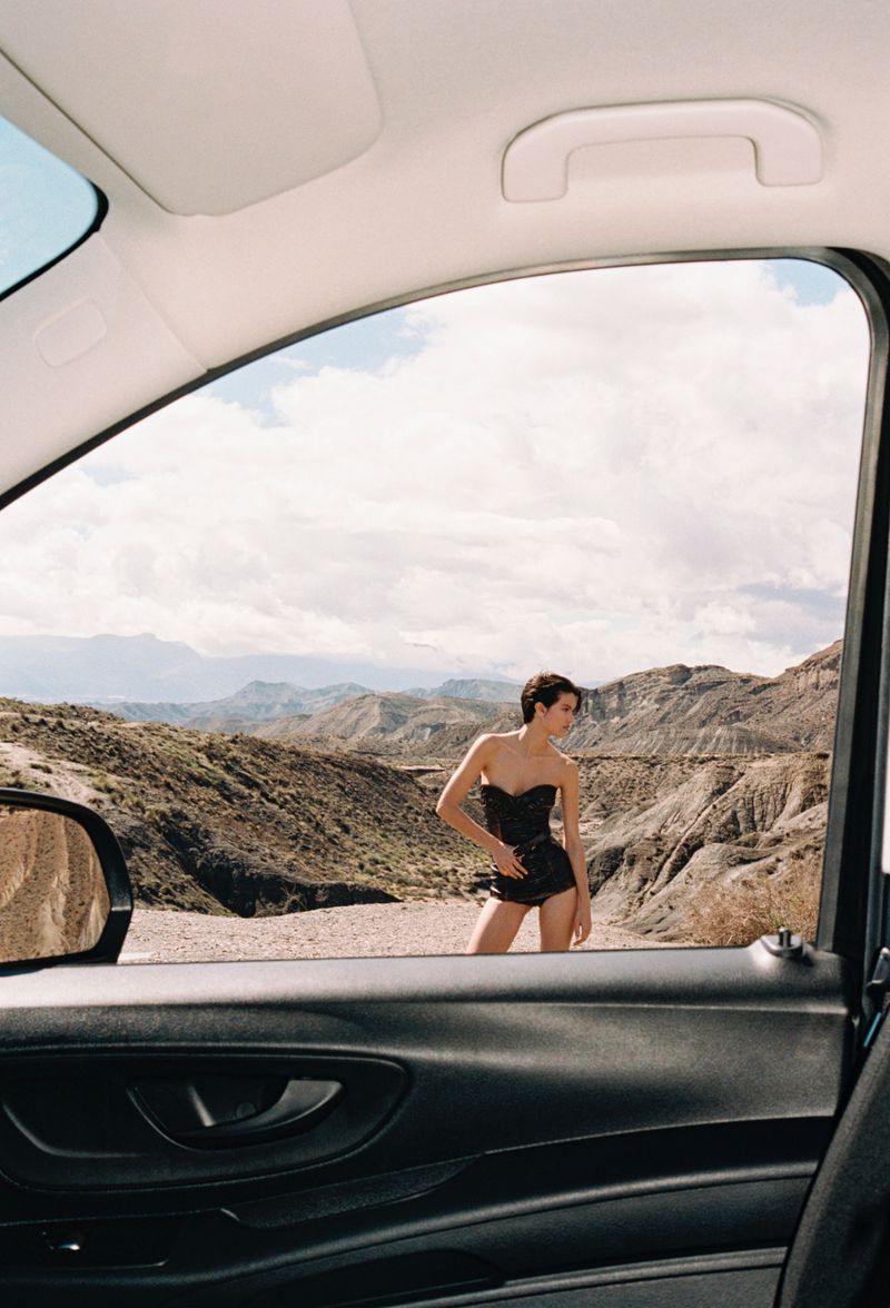 A woman in black short set poses in front of a desert mountain range, framed through the open window of a black car.