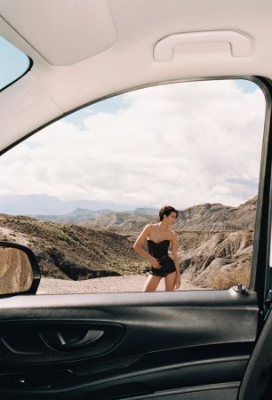 A woman in black short set poses in front of a desert mountain range, framed through the open window of a black car.
