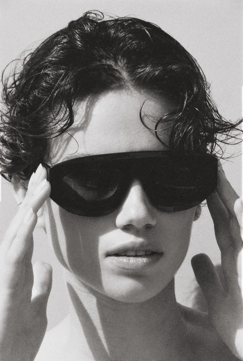 Monochrome portrait of a young adult with dark hair, wearing sunglasses and holding the frames with fingers, neutral expression.