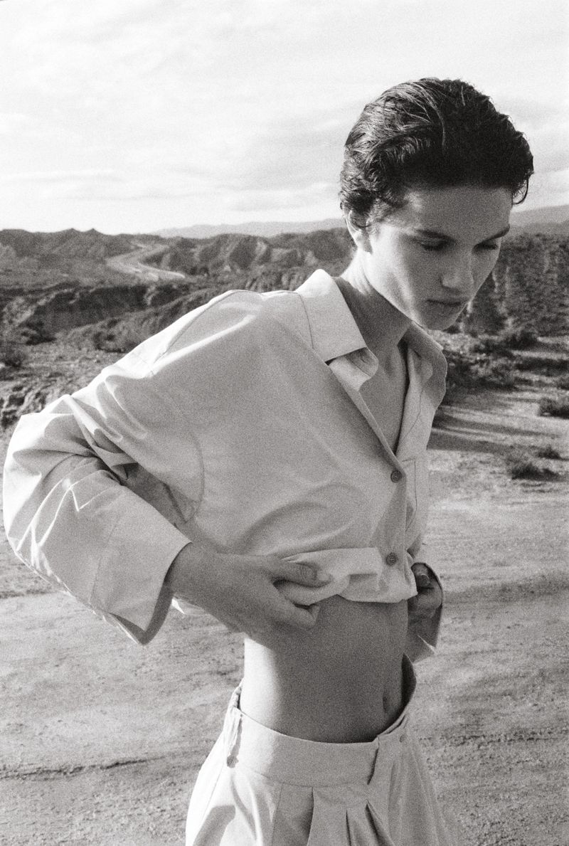 Monochrome outdoor shot of a female model in a button-down shirt and pants, revealing her midriff in a desert-like setting.