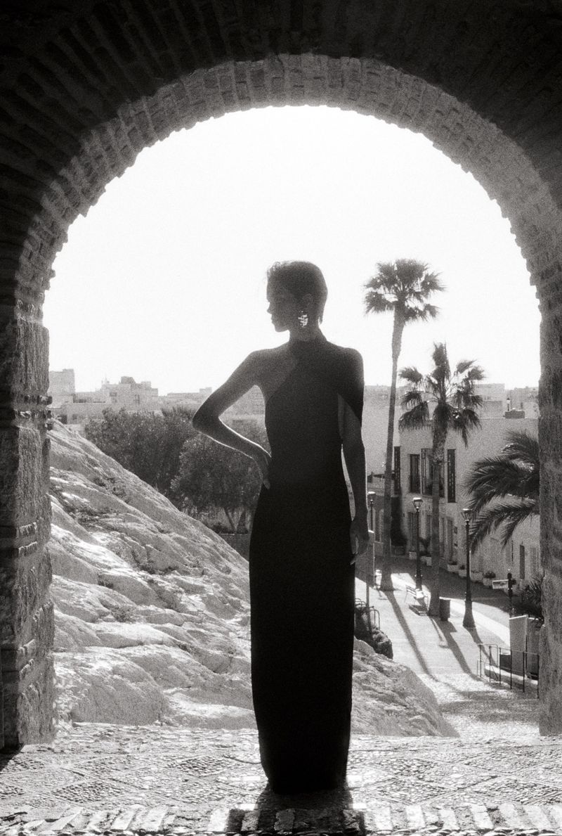Monochrome portrait of a stylish adult woman posing in an elegant dress, framed by an arched doorway with palm trees and urban backdrop.