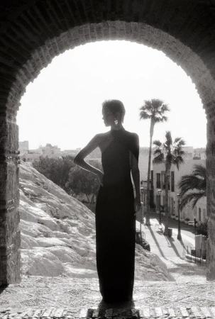 Monochrome portrait of a stylish adult woman posing in an elegant dress, framed by an arched doorway with palm trees and urban backdrop.