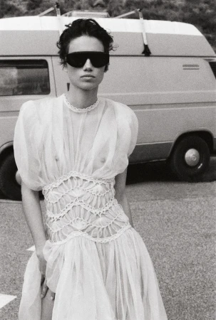 Monochrome portrait of a woman in a white dress and sunglasses, standing near a van with surfboards on top, captured in daylight.