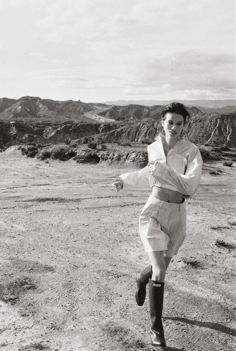 A woman in a white shirt and shorts runs across a barren landscape under a cloudy sky in a black and white outdoor fashion shot.