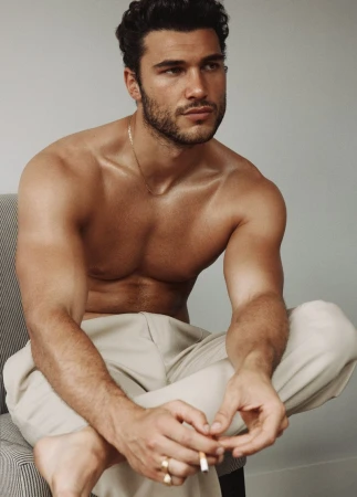 Shirtless man with tan trousers poses indoors. He has dark hair and a slight beard, seated on a chair against a grey wall in a studio.