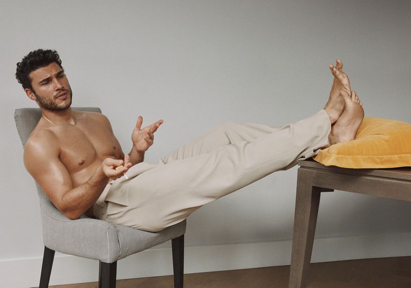 Shirtless man in light beige pants sitting in a chair, legs extended onto a small table with yellow pillow, against grey backdrop.