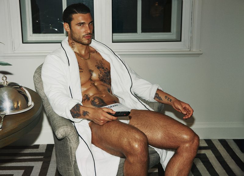 Adult male sits in an armchair wearing a white bathrobe holding a remote control in an indoor setting with a window.