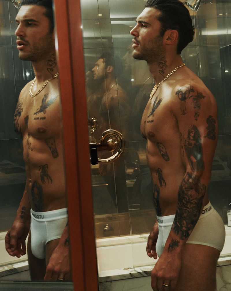 Profile view of a muscular adult male with tattoos, posing in white briefs, reflected in a mirror, inside a bathroom setting.