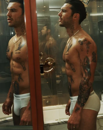 Profile view of a muscular adult male with tattoos, posing in white briefs, reflected in a mirror, inside a bathroom setting.