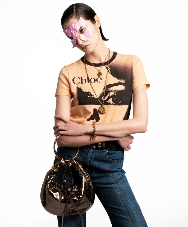 Model wearing a printed t-shirt and pink paper mask over eyes, holding a metallic handbag against a plain white background.