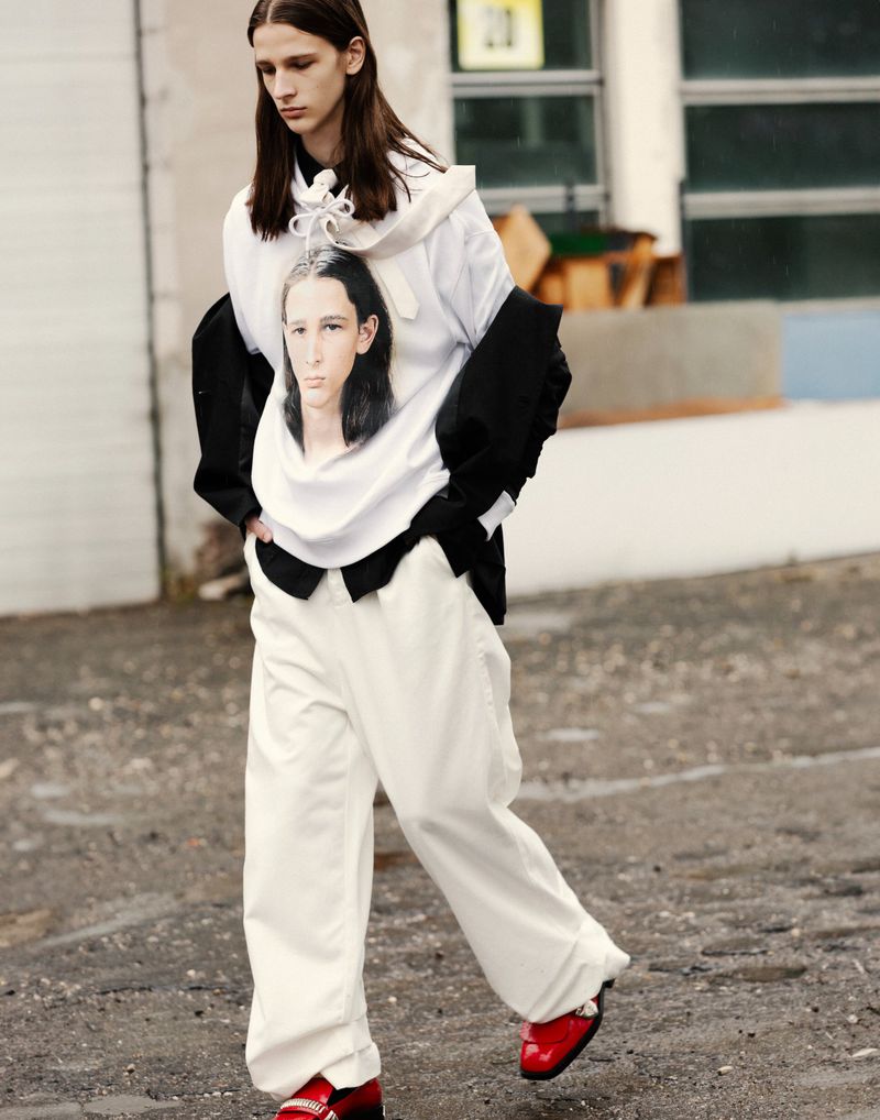 Fashion shot of a young adult in a white graphic sweatshirt, black blazer, loose white pants, and shiny red dress shoes walking on a wet street.