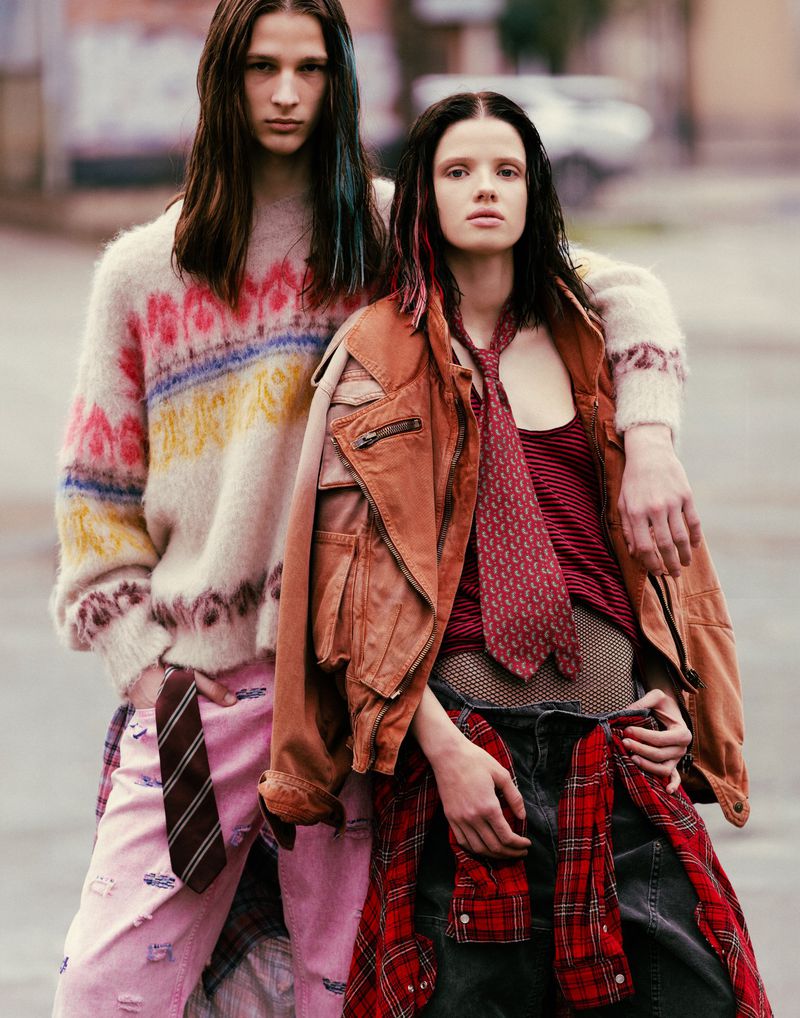 Two fashion models pose outdoors; the adult male wears a colorful sweater and the adult female wears a brown jacket and red plaid shirt.