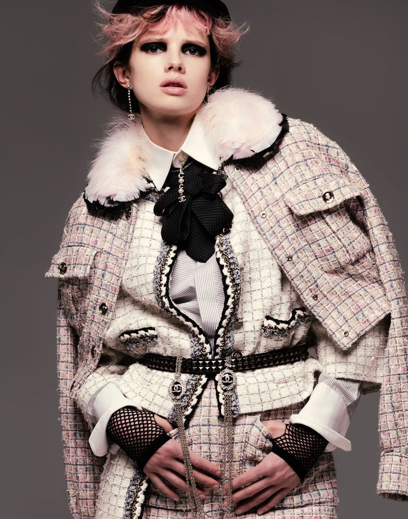 Fashionable young adult female model with pink hair wearing Chanel tweed jacket, skirt, and accessories posing indoors against gray background.