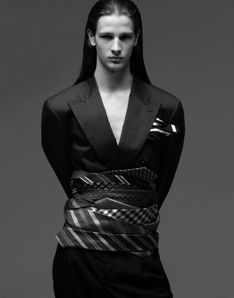 Monochrome portrait of androgynous adult model in jacket, waist wrapped with ties, standing in a studio with a neutral gray backdrop.