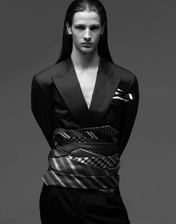 Monochrome portrait of androgynous adult model in jacket, waist wrapped with ties, standing in a studio with a neutral gray backdrop.