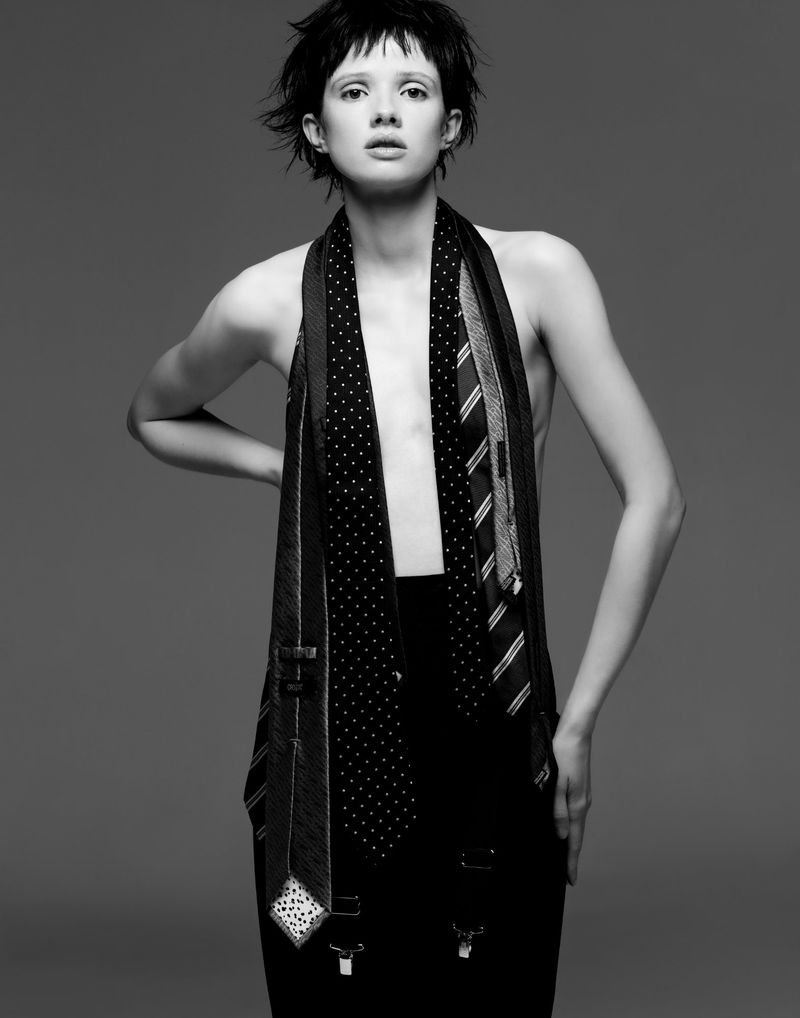 Monochrome portrait of a woman wearing ties as a garment, hands on hips, set against a plain gray background in a studio setting.