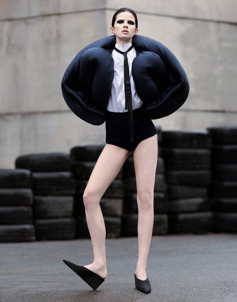 Fashion photo of a slender woman in a white collared shirt and black jacket, high-waisted briefs, and sharp-pointed flats posing indoors.