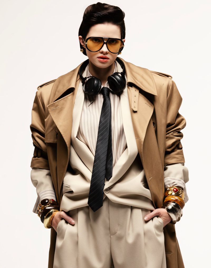 Adult female in a beige suit, trench coat, and orange aviator sunglasses posing in a studio wearing headphones and gold bangles.