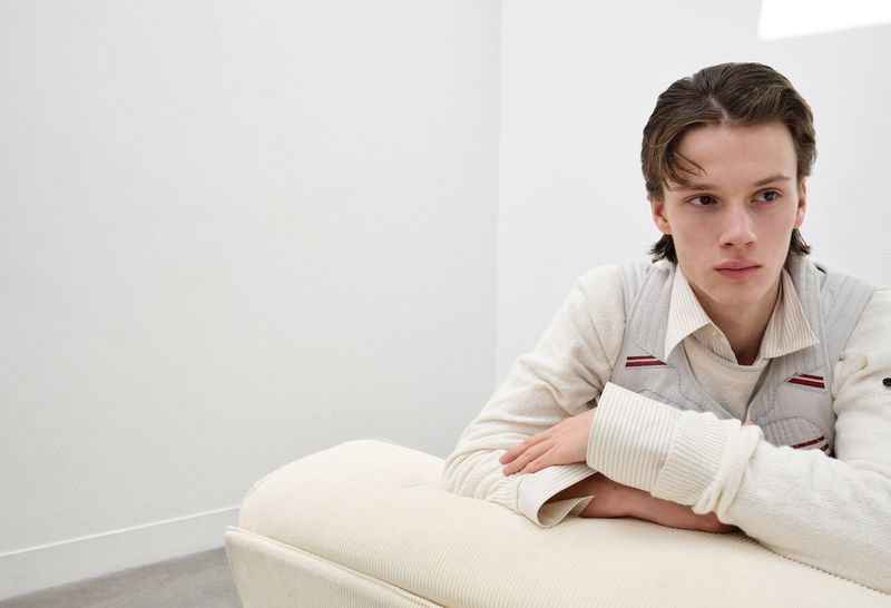Indoor portrait of a young adult man with brown hair, resting his arms on a white upholstered surface, with a clean white background.