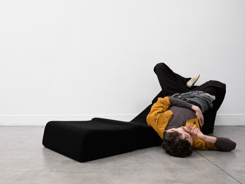 Person lying on a black modern chaise lounge in an all-white photo studio setting. The person is wearing casual clothes and light-colored shoes.