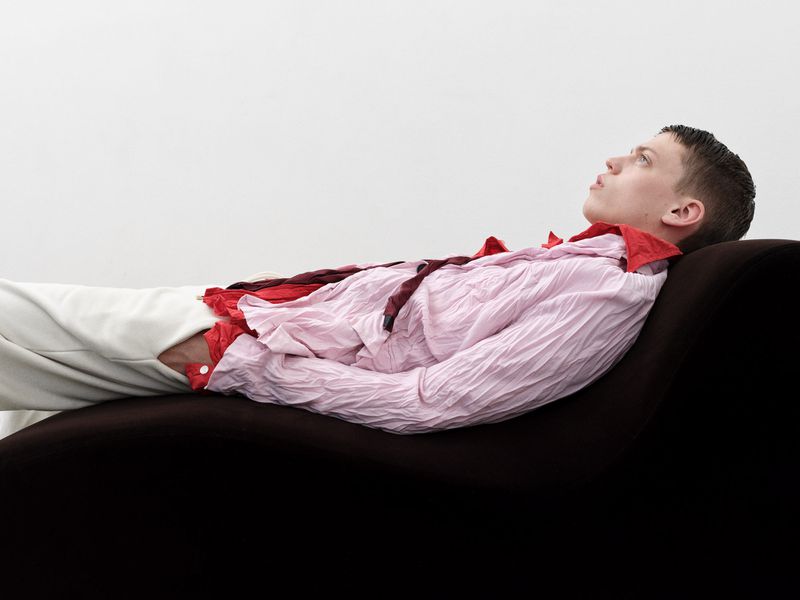 A young adult male lies on a chaise lounge, wearing a wrinkled pink and red collared shirt and white pants against a white backdrop.