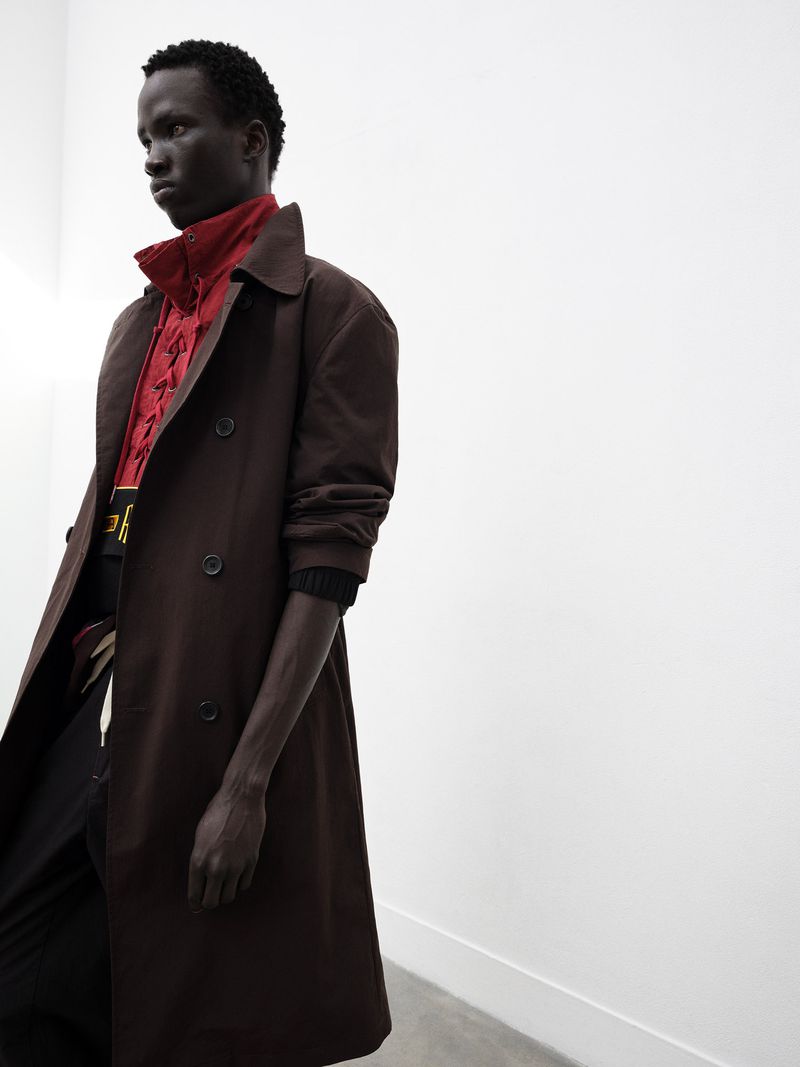 Fashionable dark-skinned adult male poses in a brown trench coat over a patterned red top indoors against a stark white wall backdrop.