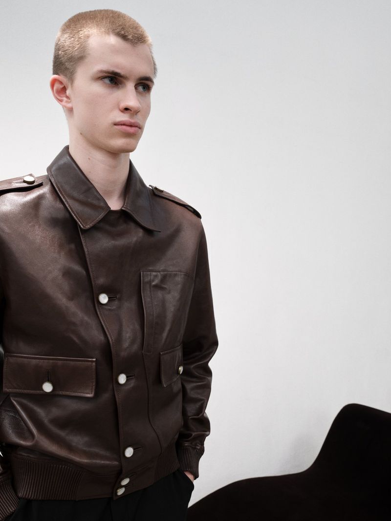 Portrait of a young fair-skinned male model in a brown leather bomber jacket with white buttons, in front of a white wall indoors.