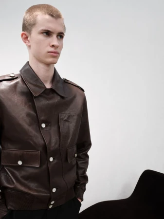 Portrait of a young fair-skinned male model in a brown leather bomber jacket with white buttons, in front of a white wall indoors.