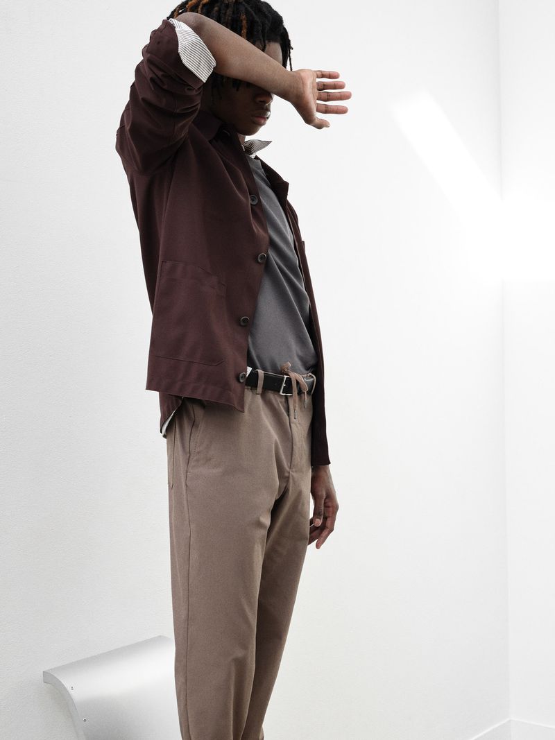 A Black person poses indoors in a brown jacket, grey shirt, and brown pants, shielding their face with a hand against a neutral wall.