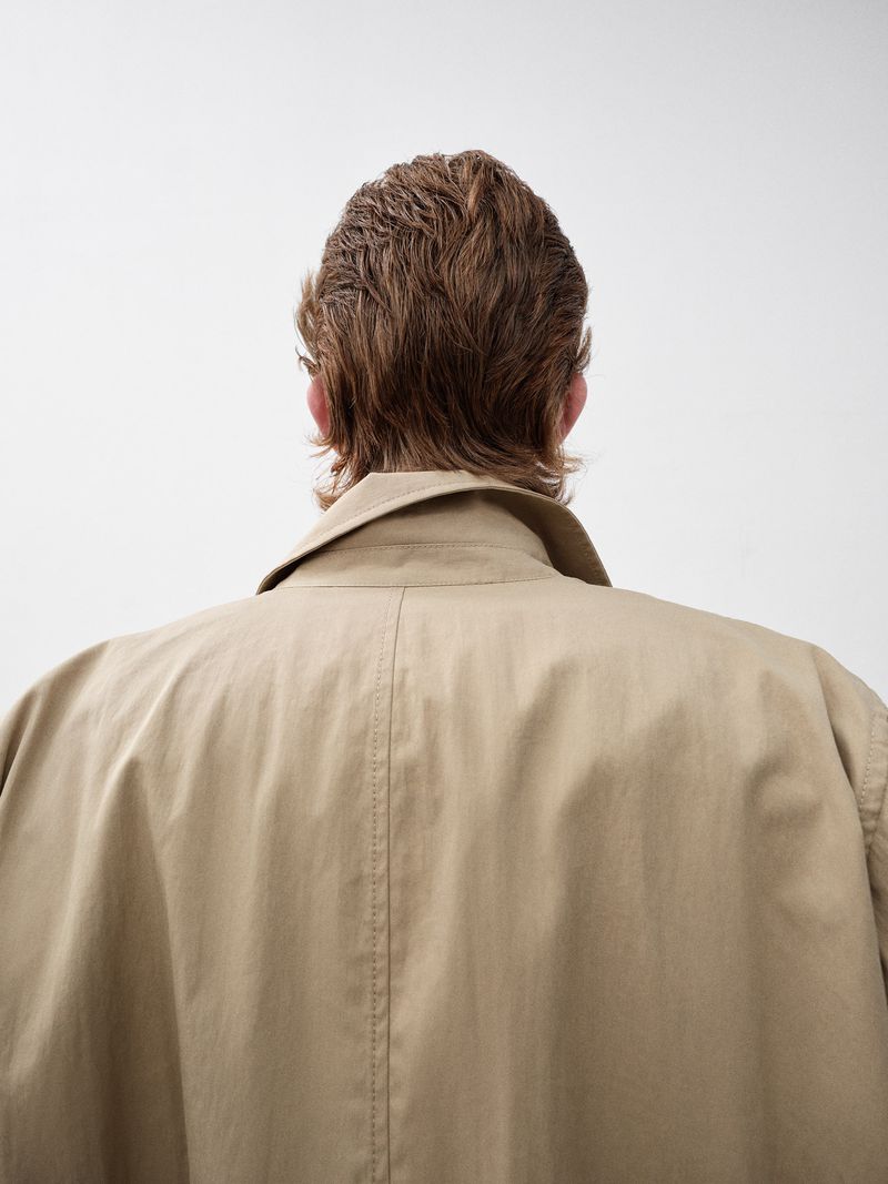Back view of a person with short brown hair wearing a beige trench coat against a white background in a studio setting.