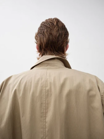 Back view of a person with short brown hair wearing a beige trench coat against a white background in a studio setting.