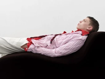 A man lying back on a curved dark lounge chair wearing a crinkled pale pink shirt and white trousers against a white background.
