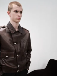 Young man with a buzz cut wearing a dark brown leather button-up jacket against a minimalist studio wall.