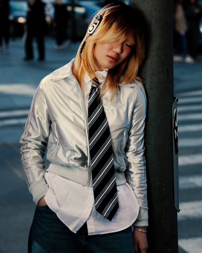 Young adult with headphones rests head on city pole in silver jacket, white shirt, tie, and jeans amidst blurred urban backdrop.
