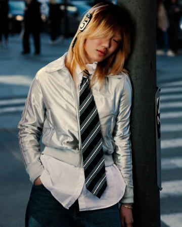 Young adult with headphones rests head on city pole in silver jacket, white shirt, tie, and jeans amidst blurred urban backdrop.