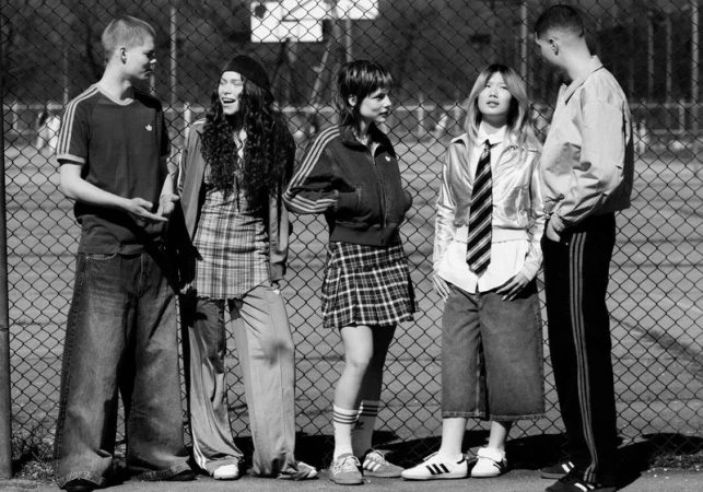 Black and white photo of five young adults standing outdoors in athletic-inspired apparel, posing against a chain-link fence in daylight.