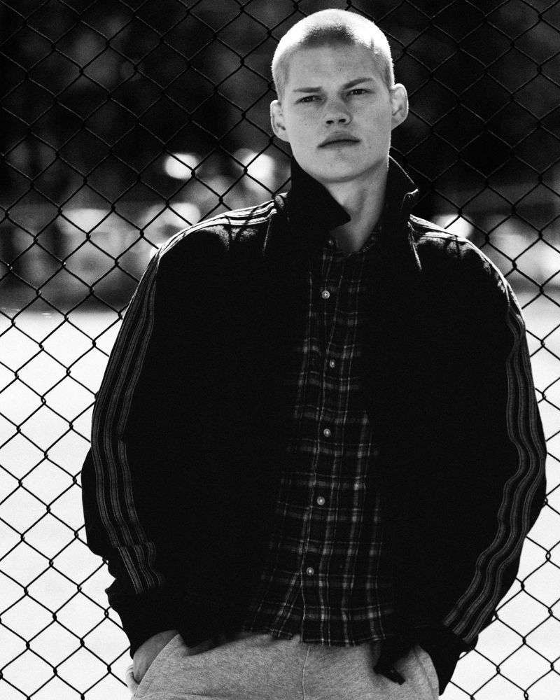 Black and white portrait of a young adult male with a shaved head wearing a jacket and shirt, posing in front of a chain-link fence.