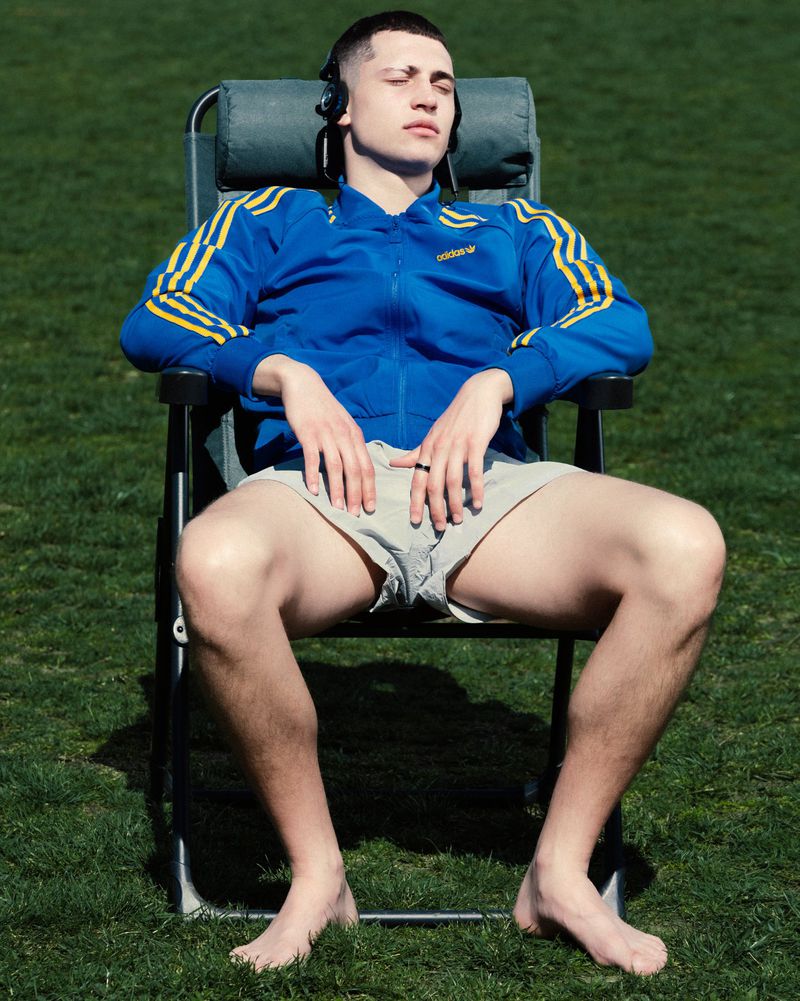 Young man relaxing in a lawn chair outdoors, wearing headphones, blue Adidas jacket, and light shorts, basking in the sun on green grass.