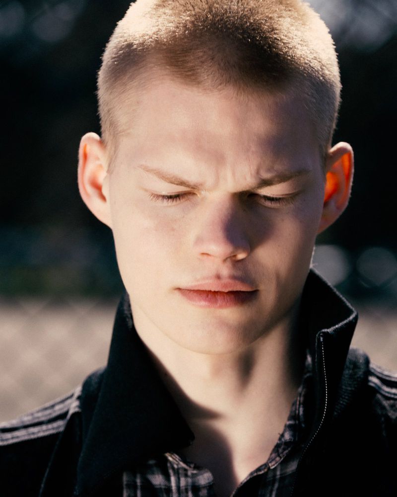Close-up portrait of a young man with a buzz cut looking down, wearing a high-collar black jacket over a plaid shirt.