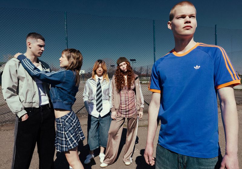 Group of five young adults outdoors in casual wear, posing against a chain-link fence with blue sky, shot in medium distance.