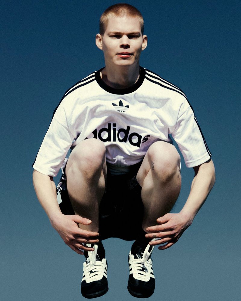 Athletic man in black-and-white Adidas shirt and shoes jumps against a vivid blue sky backdrop, showing sportswear and energy.